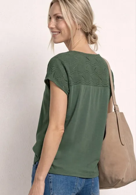 Vrouw in groen kanten shirt, blauwe jeans en beige schoudertas, haar losjes in een knot, voor een lichte muur.