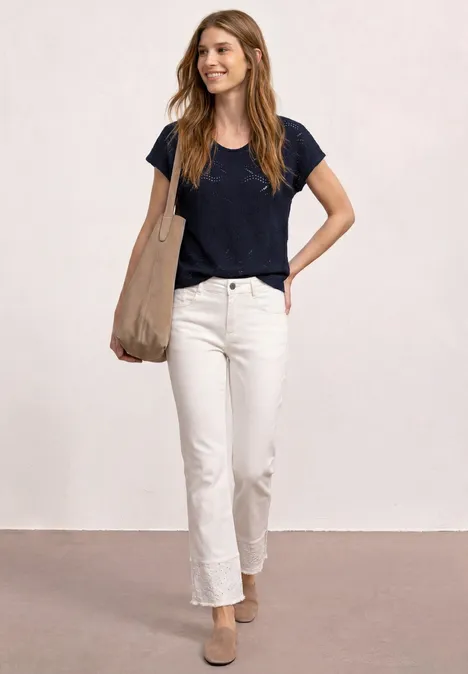 Een vrouw in een zwart t-shirt, witte jeans en beige mules draagt een grote tas tegen een lichte achtergrond.