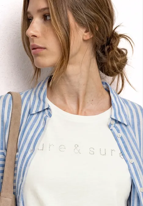 Jonge vrouw met opgestoken haar draagt wit T-shirt en blauw-wit gestreept hemd, kijkt opzij.