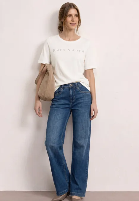 Vrouw voor neutrale achtergrond met wit T-shirt, blauwe jeans en beige schoudertas.
