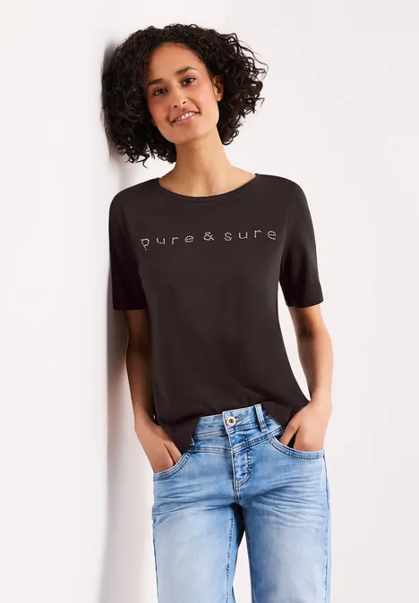 Frau lehnt an Wand, trägt schwarzen 'pure&sure' T-Shirt und blaue Jeans.
