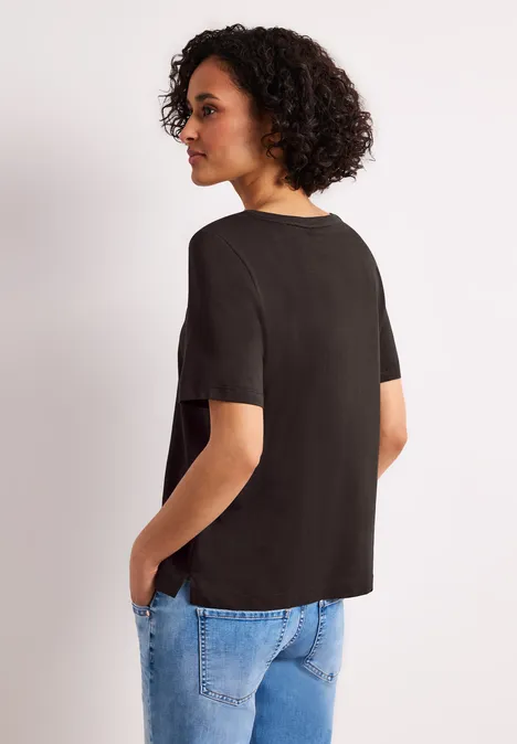 Frau mit lockigem Haar trägt schwarzes T-Shirt und blaue Jeans, steht seitlich mit Händen in den Taschen.