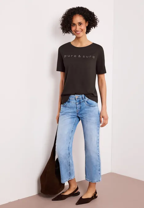 Frau in schwarzem T-Shirt mit Aufdruck, hellblauen Jeans und schwarzen Ballerinas, trägt braune Tasche