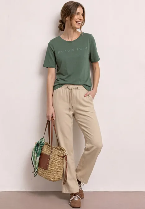 Vrouw in groen t-shirt en beige broek, staat nonchalant met rieten tas en groene sjaal.
