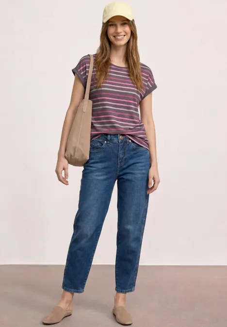 Frau steht vor hellem Hintergrund in Jeans, gestreiftem T-Shirt, flachen Schuhen und Schultertasche.