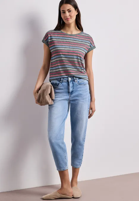 Vrouw in gestreept T-shirt, lichte jeans, houdt een trui, draagt beige instappers voor neutrale achtergrond.