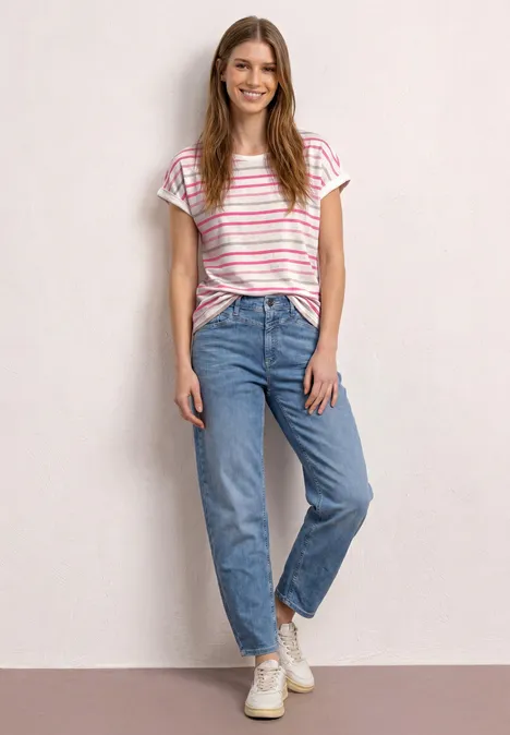 Eine Frau in gestreiftem T-Shirt, blauen Jeans und weißen Sneakern steht vor hellgrauer Wand.