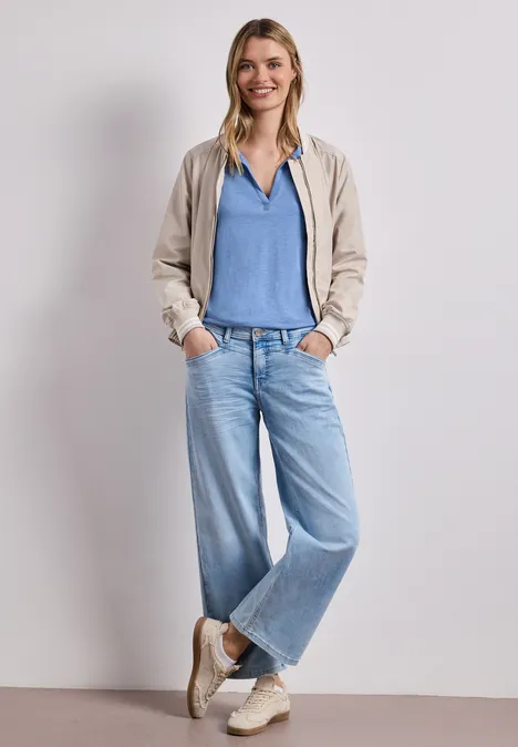 Persoon in beige jack, blauw shirt, wijde spijkerbroek en sneakers, ontspannen met handen in de zakken.