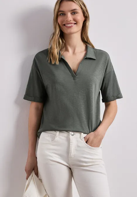 Vrouw draagt olijfgroene polo met korte mouwen en lichte, hoge jeans, houdt een tas vast.