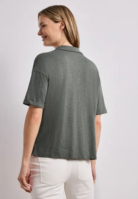 Rugaanzicht van een vrouw in een donker groen shirt en lichte broek voor een lichte achtergrond.