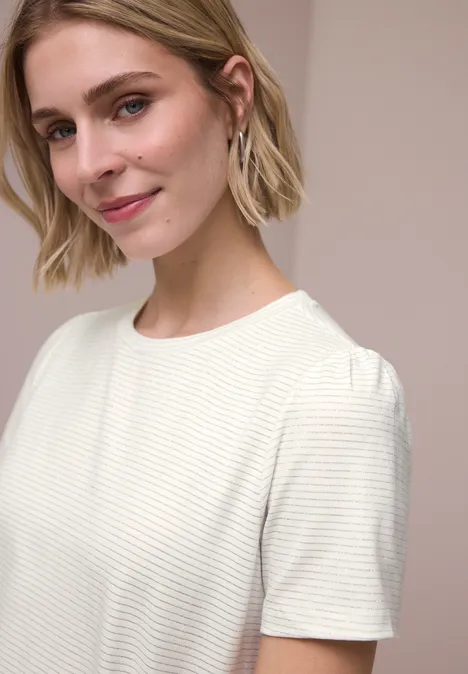 Vrouw met kort blond haar in een wit geribd T-shirt met korte mouwen en oorbellen voor een neutrale achtergrond.