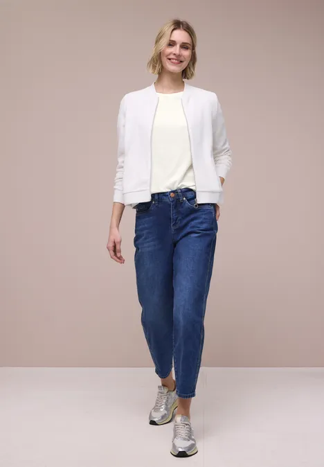 Een vrouw draagt een witte jas, licht T-shirt, blauwe jeans en zilveren schoenen voor een beige achtergrond.