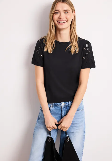 Shirt met ronde hals en kanten mouwen Black Vrouw in zwarte T-shirt en lichtblauwe jeans staat en houdt een tas voor haar lichaam vast.