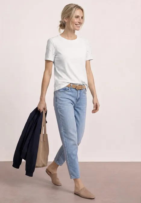Vrouw in wit T-shirt, blauwe spijkerbroek en beige schoenen, met donkerblauwe jas en tas in de hand.