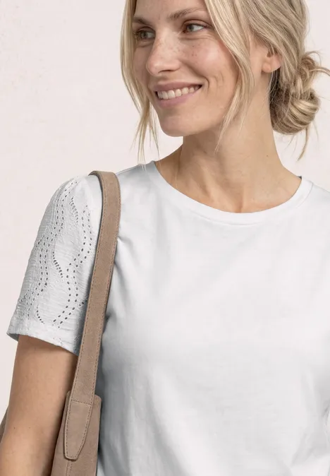 Glunderende vrouw in wit t-shirt met gehaakte mouwen en schoudertas voor een lichte achtergrond.