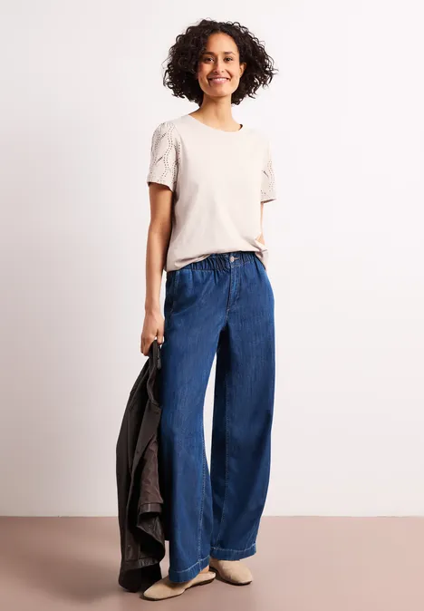 Shirt met ronde hals en kanten mouwen clay sand Vrouw met beige t-shirt en wijde blauwe jeans, houdt een zwart jack vast, lichte schoenen, neutrale achtergrond.