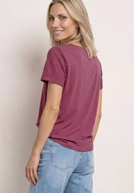 Shirt met ronde hals en kanten mouwen dark clouded blush Persoon met halflang blond haar zijwaarts voor een lichte achtergrond, draagt frambooskleurig T-shirt en blauwe spijkerbroek.