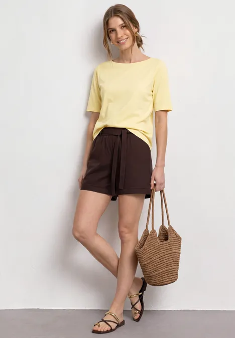 Basic T-Shirt mit U-Boot-Ausschnitt soft yellow Person in gelbem T-Shirt, braunen Shorts, mit Basttasche und schwarzen Sandalen an Wand.