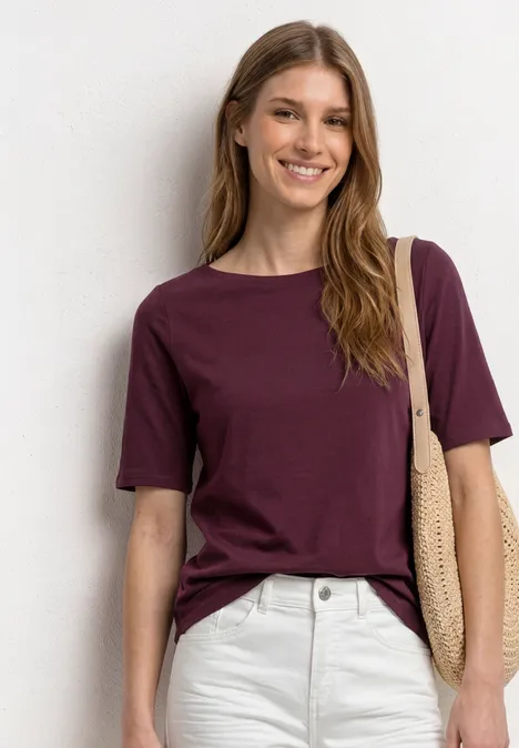 Basic T-Shirt mit U-Boot-Ausschnitt rustic rouge Eine Frau trägt ein dunkelviolettes T-Shirt, weiße Hose und eine Strohtasche.