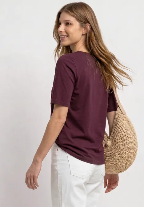 Basic T-Shirt mit U-Boot-Ausschnitt rustic rouge Frau mit weinrotem T-Shirt und weißer Hose trägt eine runde Strohtasche über der Schulter.