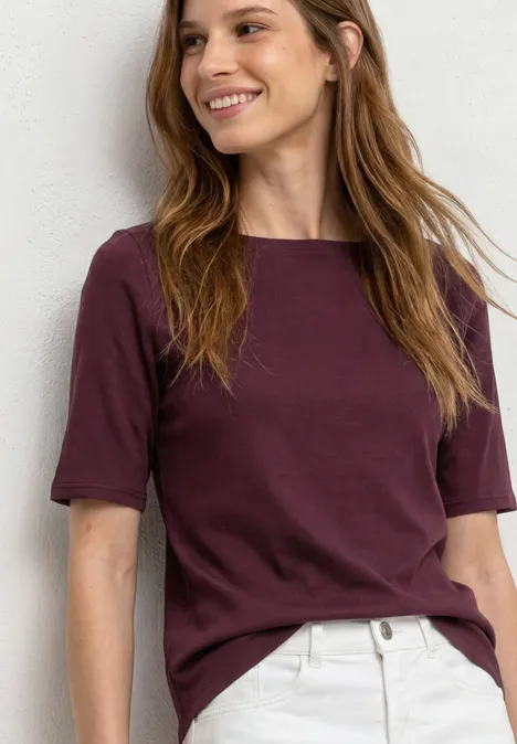 Basic T-Shirt mit U-Boot-Ausschnitt rustic rouge Frau in bordeauxfarbenem T-Shirt und hellbeiger Hose vor weißer Wand.
