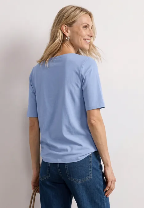 Basic T-Shirt mit U-Boot-Ausschnitt northern blue Frau von hinten mit hellblauem T-Shirt und Jeans steht neben einem weißen Hocker in weißem Raum.