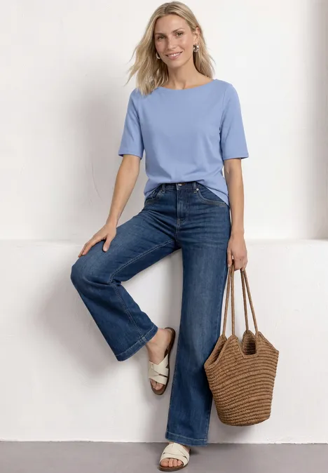 Basic T-Shirt mit U-Boot-Ausschnitt northern blue Frau in hellblauem T-Shirt, ausgestellten Jeans und Basttasche vor weißem Hintergrund.