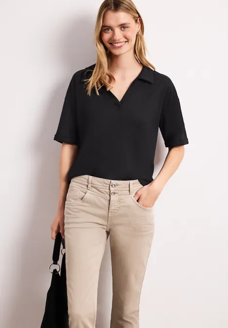 Een vrouw draagt een zwart overhemd met V-hals en beige broek, en houdt een klein zwart tasje.