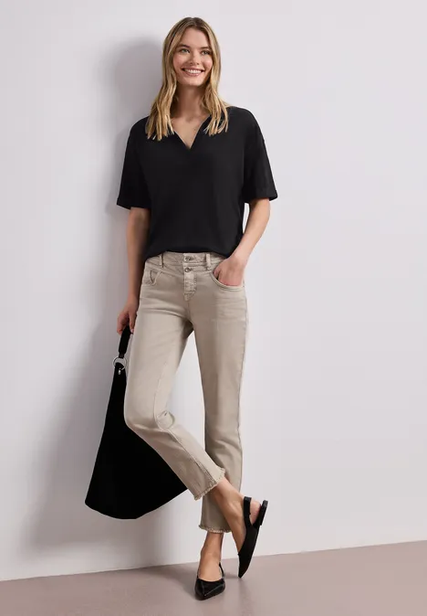 Vrouw in zwart shirt en beige broek met zwarte tas leunt tegen een witte muur.