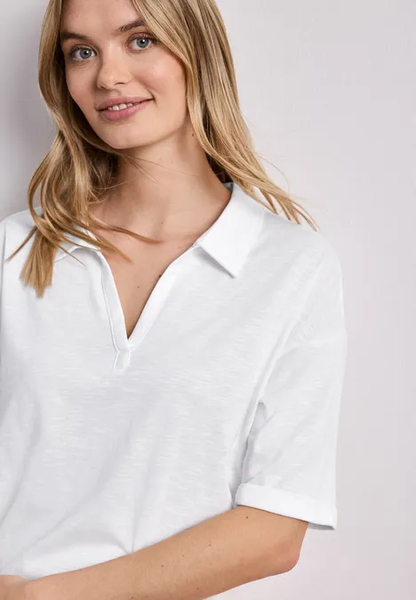 Junge Frau lächelt in schlichtem weißen Poloshirt, vor neutralem Hintergrund.