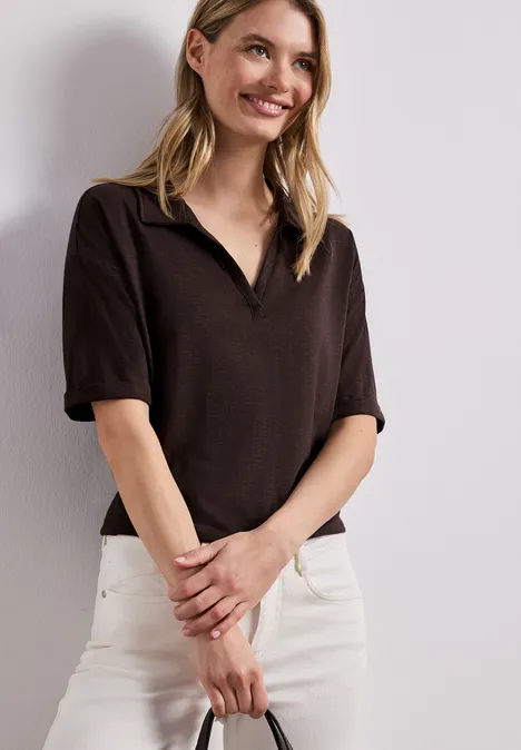 Person vor heller Wand, trägt braunes kurzärmeliges Poloshirt und weiße Jeans, Hände vorn verschränkt.