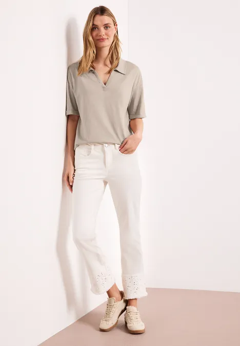 Vrouw tegen witte muur met grijs poloshirt, witte jeans met kanten zoom en lichte sneakers.