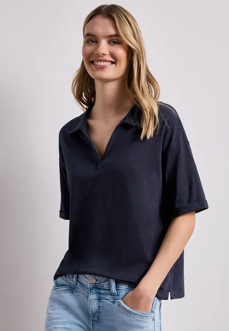 Persoon voor lichte muur, draagt losvallend donkerblauw poloshirt, één hand in de spijkerbroekzak.