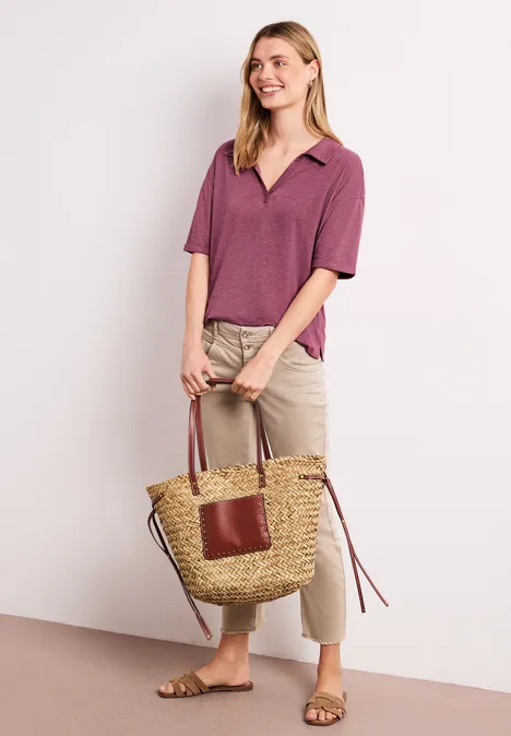 Vrouw in paars polo, beige broek en sandalen met een gevlochten tas voor een lichte achtergrond.