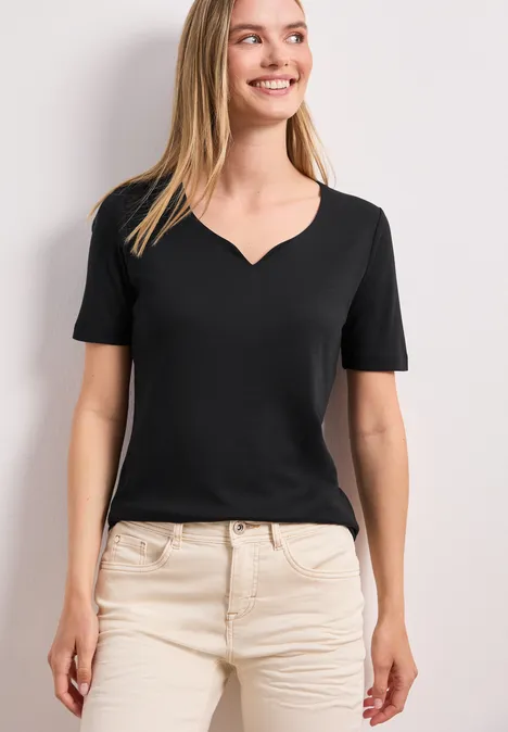 Frau in schwarzem V-Neck-T-Shirt und beiger Hose vor weißem Hintergrund.