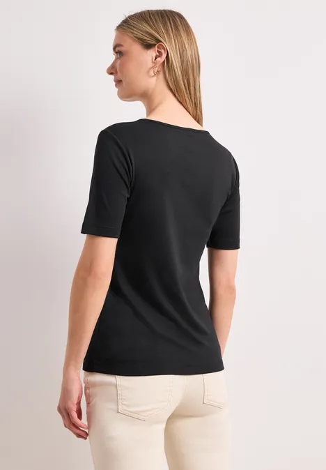 Blonde Frau von hinten in schwarzem T-Shirt und beigen Hosen vor hellem Hintergrund.