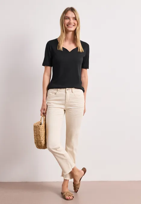Frau in schwarzem V-Shirt, hellen Jeans und braunen Mokassins mit Korbtasche vor neutralem Hintergrund.