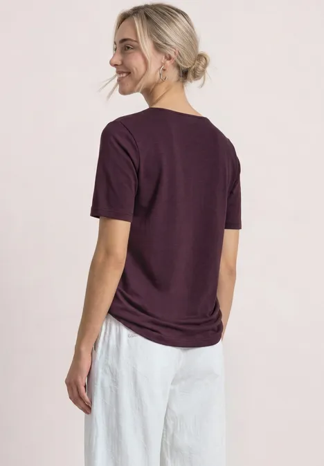 Eine Frau von hinten mit lila T-Shirt und weißer Hose, die lächelnd zur Seite schaut.