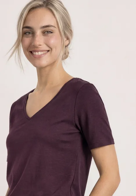 Frau mit hellblonden Haaren lächelt in violettem V-Ausschnitt-T-Shirt vor weißem Hintergrund.