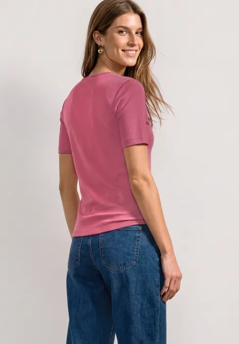Eine Frau in einem pinken T-Shirt und blauen Jeans steht seitlich mit Blick über die Schulter.