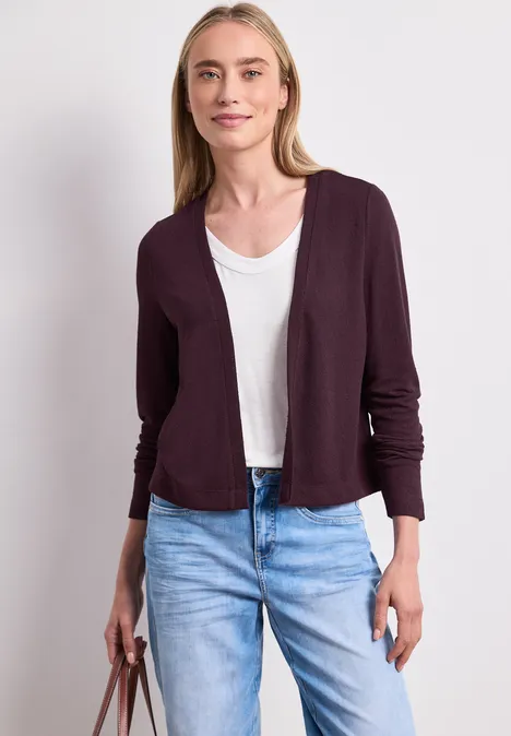 Frau in weißem T-Shirt, offener dunkelroter Strickjacke, hellen Jeans und mit einer braunen Handtasche.