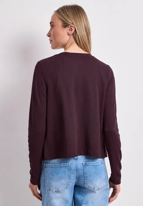 Frau von hinten in dunkellila Langarmshirt und hellblauen Jeans vor neutralem Hintergrund.