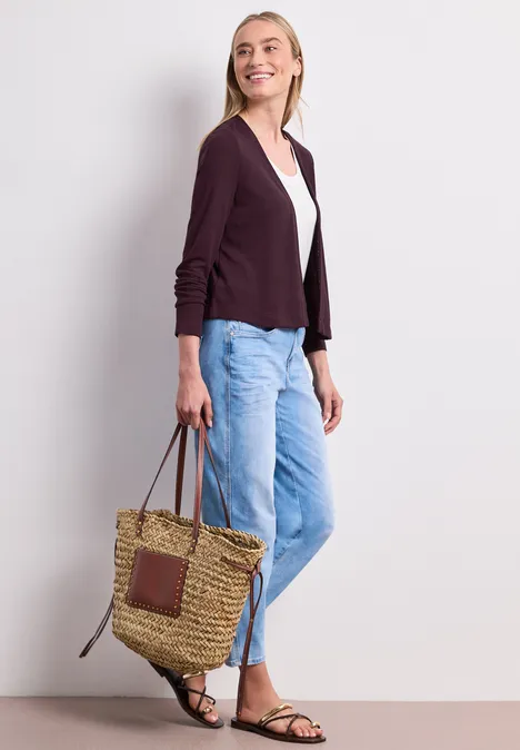 Frau in lässigem Outfit mit Cardigan, weißem Shirt, blauen Jeans, Sandalen und geflochtener Tasche.