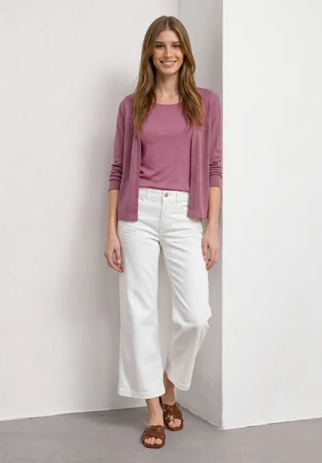 Femme en pull rose, jean culotte blanc et sandales marron, posant devant un mur blanc.