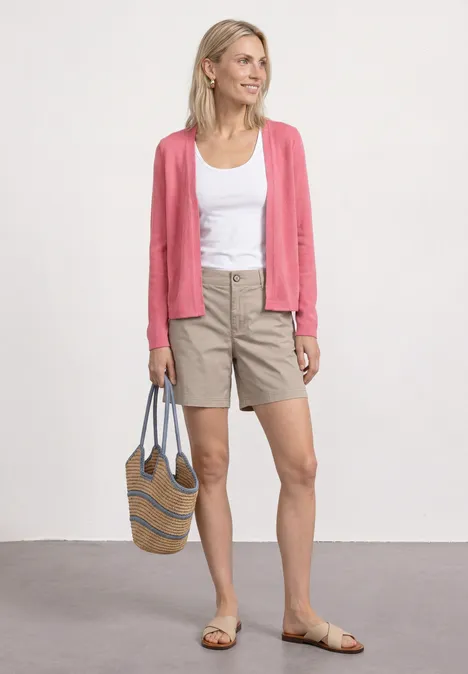 Femme en débardeur blanc, cardigan rose, short beige et sandales, tenant un sac.