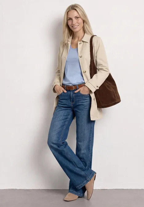 Femme appuyée sur un mur blanc, portant un trench beige, un top bleu clair, un jean, ballerines beiges et un grand sac.