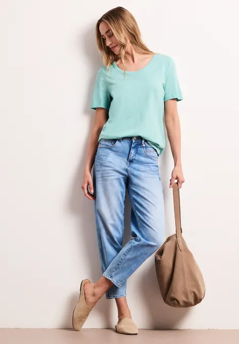 Vrouw leunt tegen een muur, draagt een mintgroen shirt, losse jeans, beige espadrilles en een grote tas.