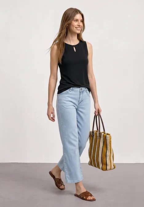 Een vrouw draagt een zwart topje, lichte jeans, bruine sandalen en houdt een gestreepte tas vast.