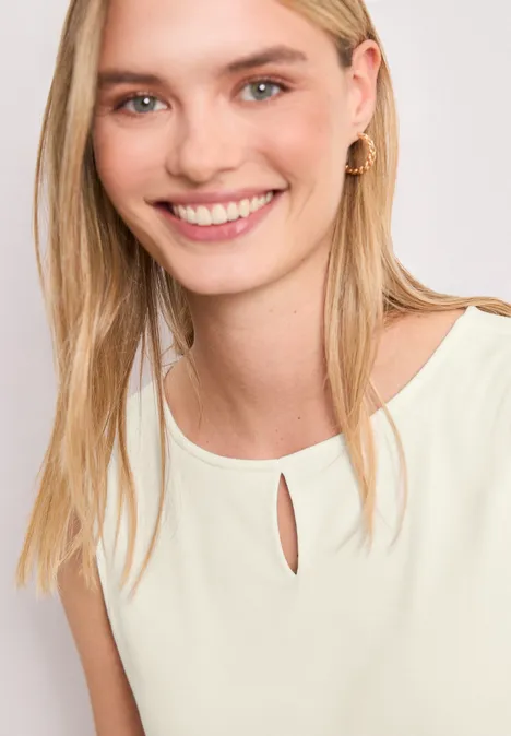 Vrouw met lang blond haar draagt een mouwloze, lichtcrèmekleurige top met een klein sleutelgatdetail bij de hals.