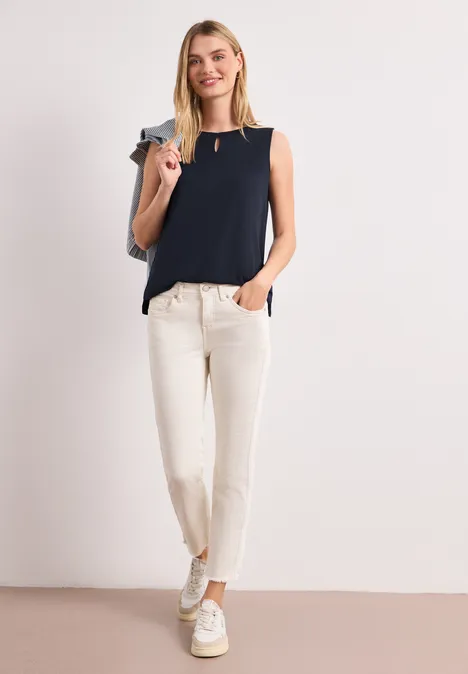 Eine Frau trägt ein ärmelloses dunkelblaues Top, helle Hose und weiße Sneaker, mit Tasche über der Schulter.